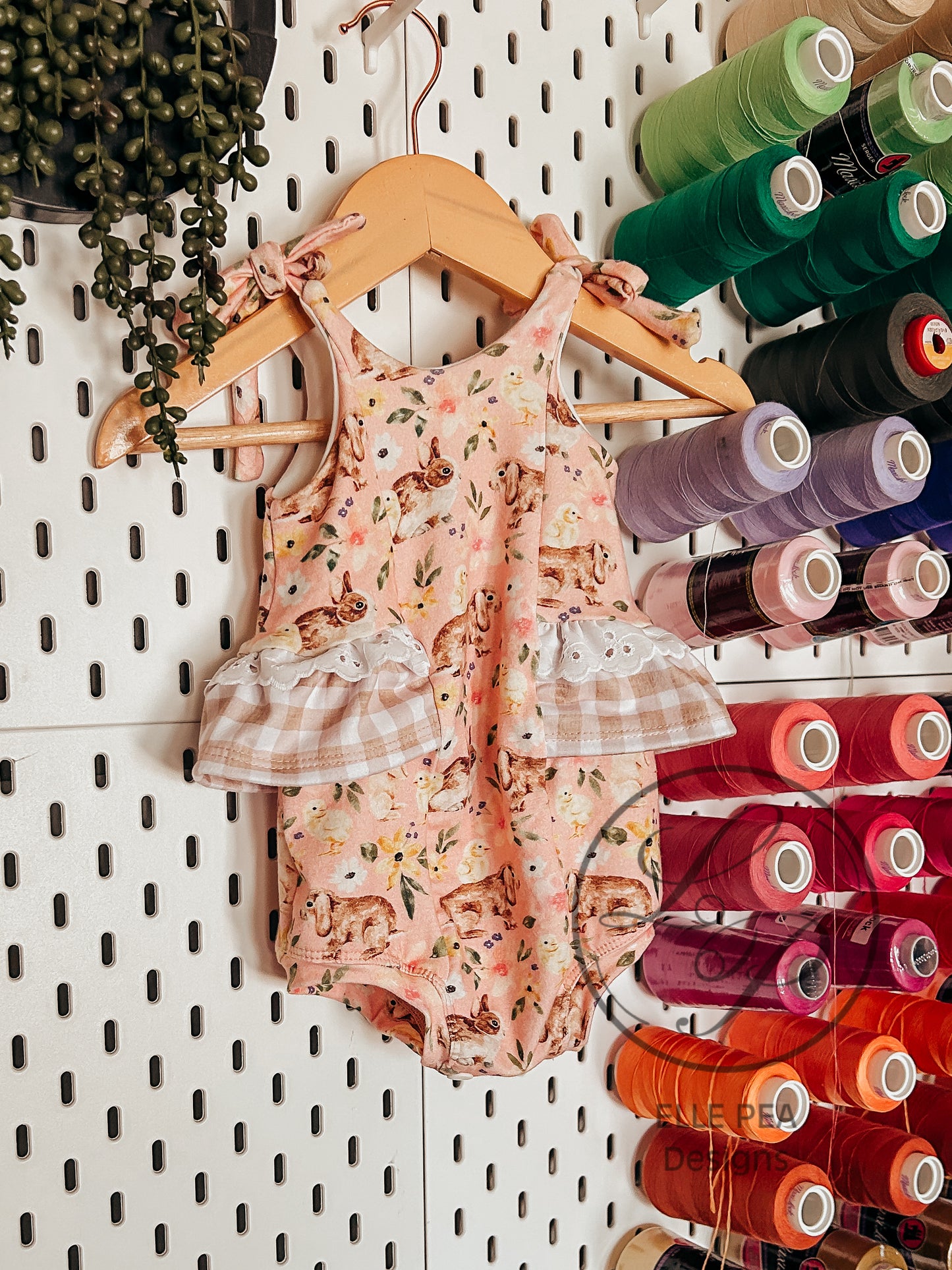 Children's floral romper on a wooden hanger against a pegboard with spools of thread.