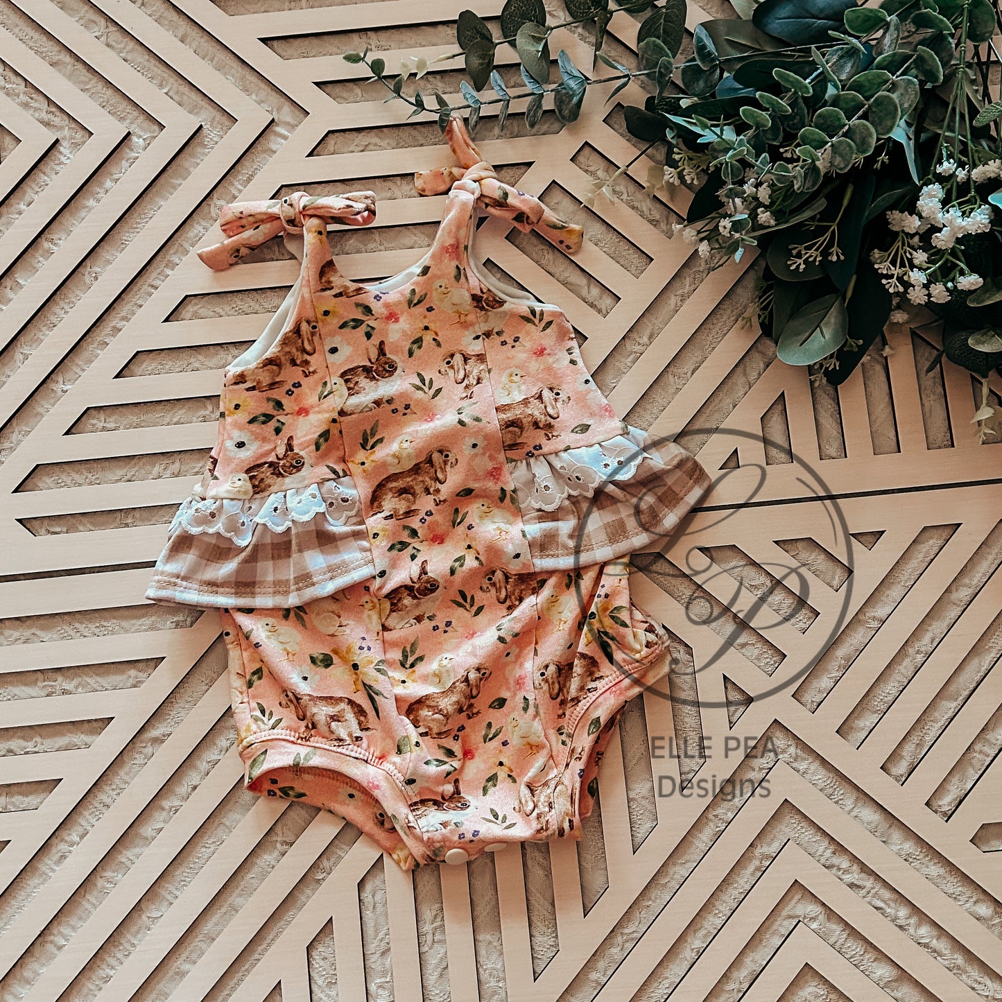 Floral baby romper with plaid accents on a geometric wooden surface with greenery.