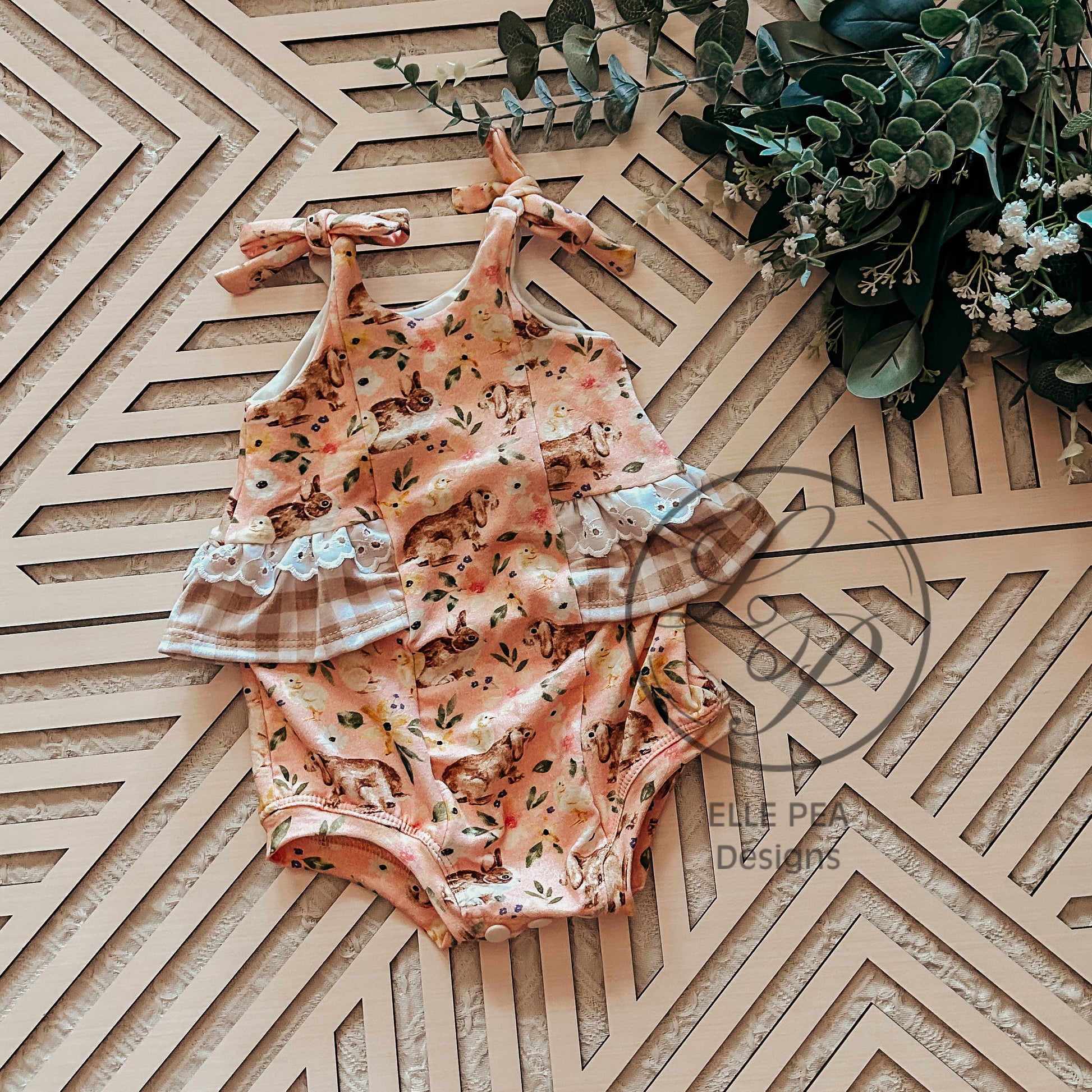 Floral baby romper with plaid accents on a geometric wooden surface with greenery.